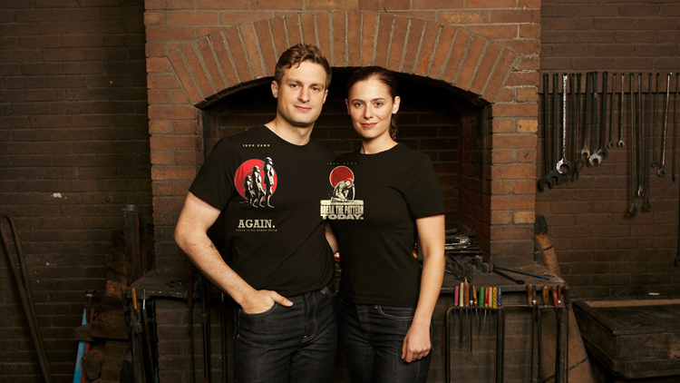 Two people in black Iron Dawn tees in a blacksmith's forge