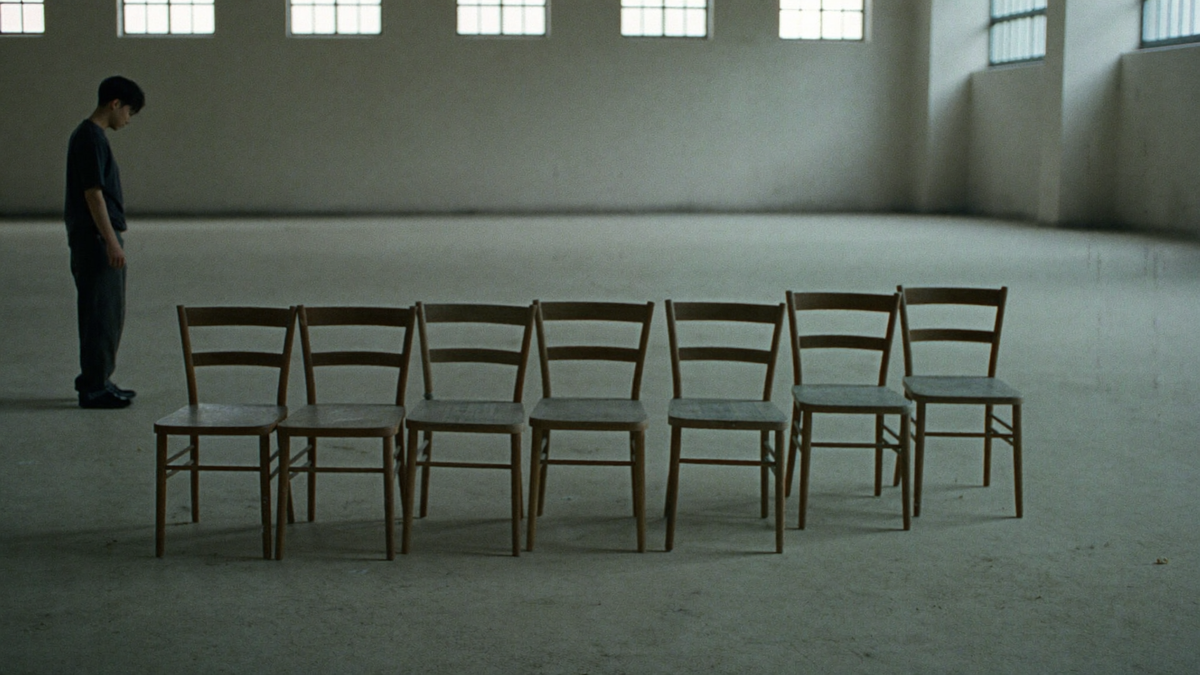 Rows of empty wooden chairs in a bare hall lit from high windows
