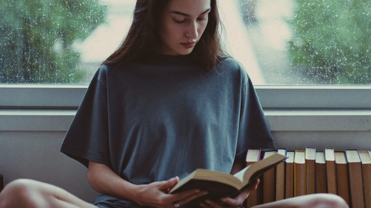 A reader curled into a window seat with rain on the glass