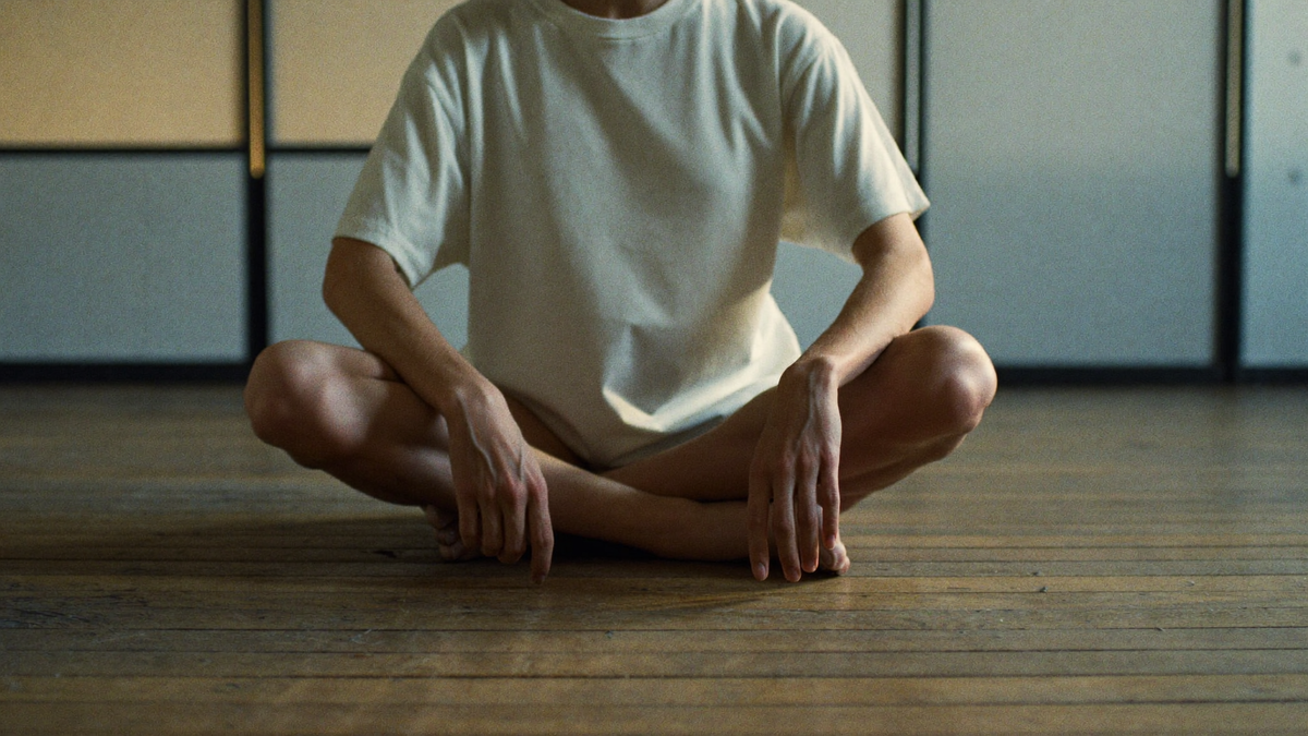 A figure sitting still on a worn wooden floor in a spare empty hall