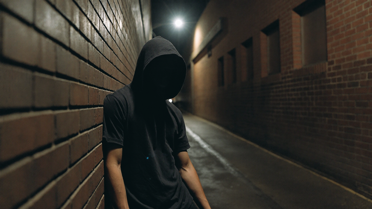 A hooded figure against a wet brick wall in a dark alley