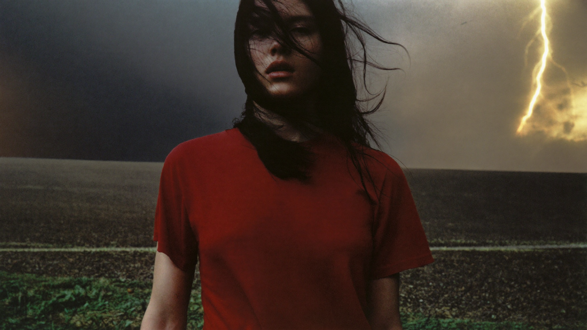 A figure in a field under a violent storm sky, wind pulling at their clothes