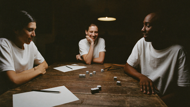 Three friends around a scarred table under one low lamp, dice scattered