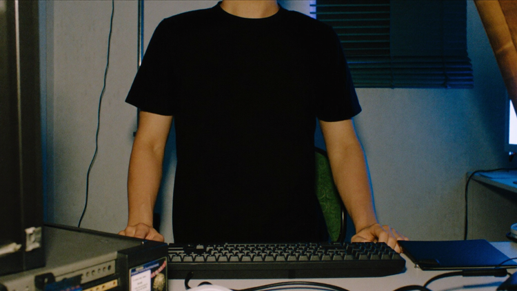 A dim home office lit by monitor glow, mechanical keyboard, plain black tee